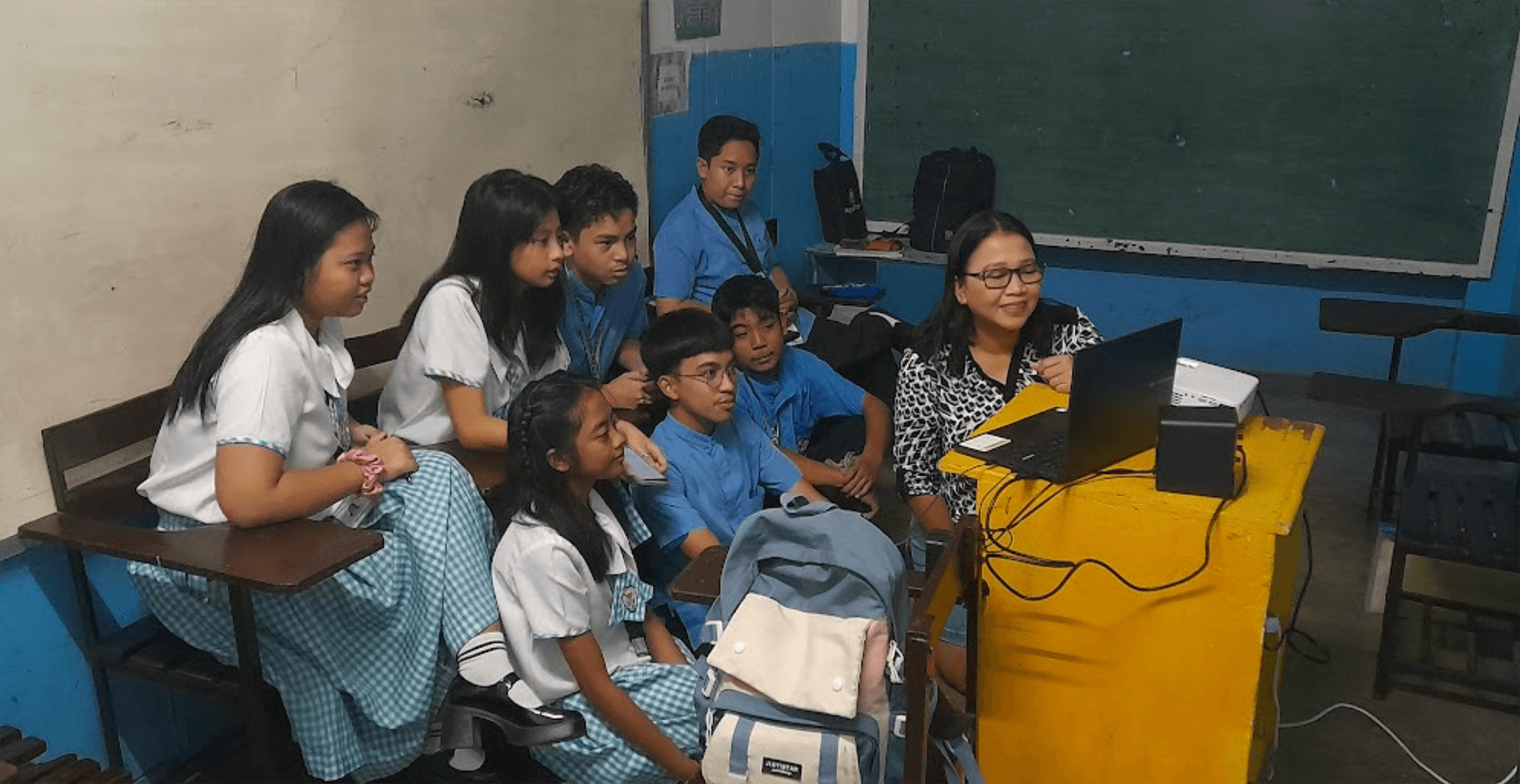 Teacher working at a laptop with several students around her looking at the screen.